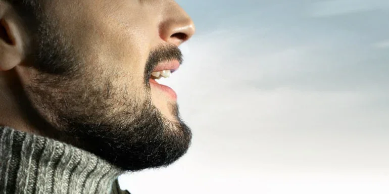 Profile of a man with a beard and open mouth, wearing a grey sweater, against a light blue sky.